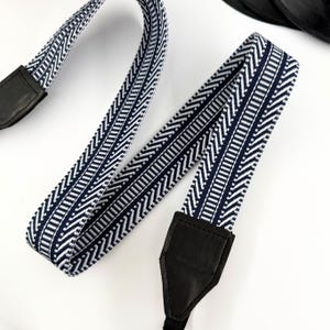 Puede incluir: Una correa de cámara con estampado azul marino y blanco con extremos de cuero negro. La correa presenta un diseño geométrico repetitivo. La correa está diseñada para usarse alrededor del cuello o el hombro.