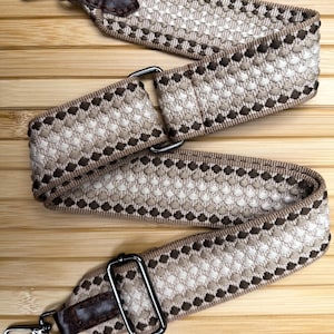 May include: A wide, adjustable bag strap featuring a woven geometric pattern in shades of brown, beige, and cream. The strap includes dark brown faux leather end tabs, dark metal swivel lobster clasps, and a rectangular metal sliding buckle.