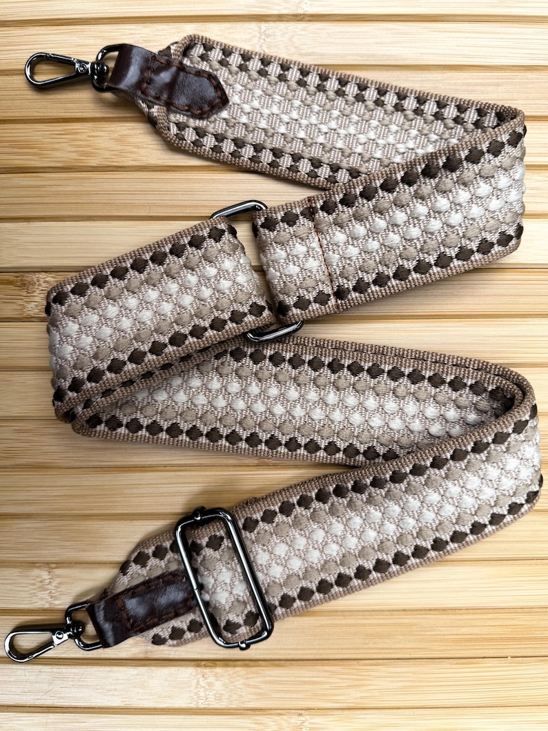 May include: A wide, adjustable bag strap featuring a woven geometric pattern in shades of brown, beige, and cream. The strap includes dark brown faux leather end tabs, dark metal swivel lobster clasps, and a rectangular metal sliding buckle.