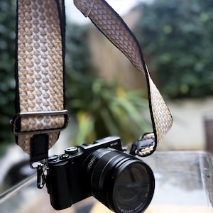 May include: A black digital camera with a lens attached to a woven camera strap. The strap features a repeating geometric pattern in shades of beige, white, and brown, with black leather ends and dark metal hardware for adjustable length.