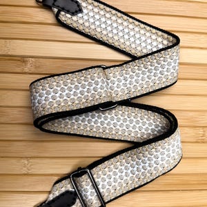 May include: A woven bag strap featuring a repeating geometric pattern in beige, white, and black. The strap includes black leather end tabs with visible stitching, dark metal swivel lobster clasps, and an adjustable metal slider buckle.
