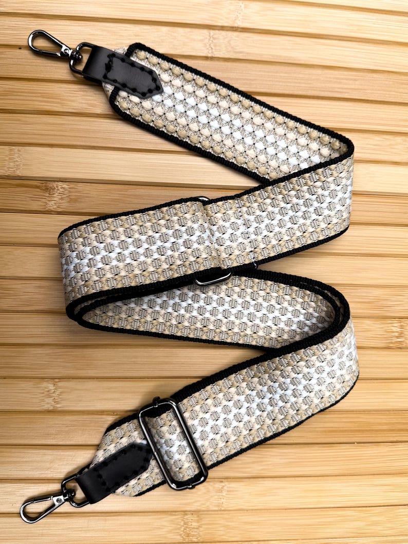 May include: A woven bag strap featuring a repeating geometric pattern in beige, white, and black. The strap includes black leather end tabs with visible stitching, dark metal swivel lobster clasps, and an adjustable metal slider buckle.
