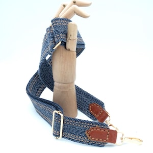 Puede incluir: Una correa de bolso tejida azul y beige con herrajes metálicos dorados y detalles de cuero marrón. La correa está sujeta a una mano de maniquí de madera, mostrando su diseño. La correa presenta un patrón repetitivo.