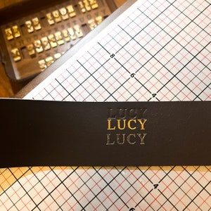 May include: A dark brown leather strip featuring the name LUCY embossed three times in a vertical column. The top instance is a blind deboss, the middle is gold foil stamped, and the bottom is a silver foil stamp, positioned over a grid mat.