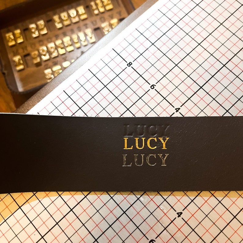 May include: A dark brown leather strip featuring the name LUCY embossed three times in a vertical column. The top instance is a blind deboss, the middle is gold foil stamped, and the bottom is a silver foil stamp, positioned over a grid mat.