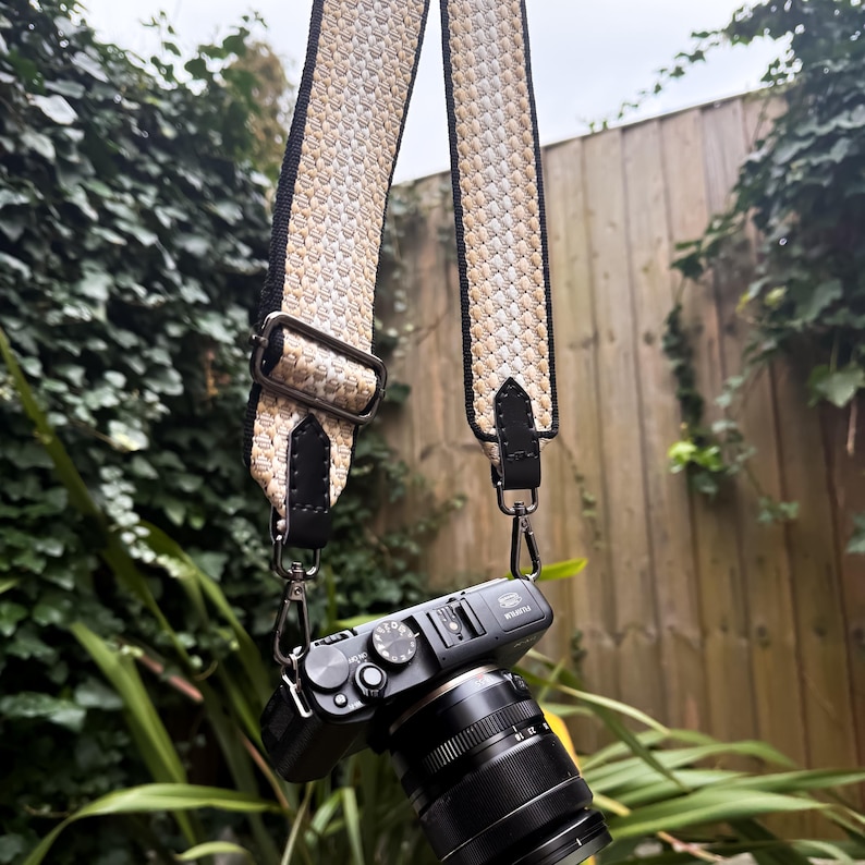 May include: A black digital camera with a lens attached, featuring a wide, adjustable camera strap. The strap has a woven, textured pattern in shades of beige, cream, and white, bordered by black edges with black leather tabs and metal hardware.