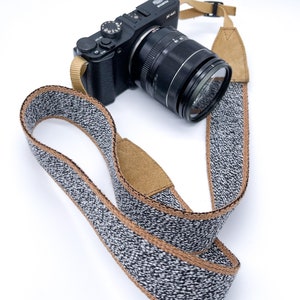 Puede incluir: Una cámara negra con una lente y una correa de cámara gris y marrón. La cámara tiene un dial plateado y el texto "FUJIFILM X-M1" en la parte superior.