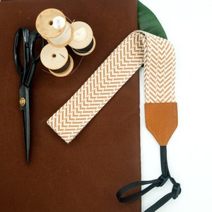 Pode incluir: Uma correia de câmera com padrão chevron marrom e branco com uma correia ajustável de couro marrom. A correia está sobre uma superfície de tecido marrom com carretéis de fio.