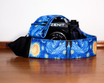May include: A black Zenit camera with a camera strap featuring a blue, yellow, and white swirling pattern reminiscent of a starry night sky. The strap has black fabric ends and is draped around the camera body, showcasing the detailed print.