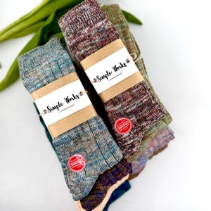 Könnte beinhalten: Mehrere gestapelte, farbenfrohe Socken, jede mit einem braunen Papierband mit der Aufschrift "Simple Works" umwickelt. Die Socken sind in Blau-, Grün- und Brauntönen gehalten, mit einem roten "Organic"-Aufkleber. Grüne Blätter sind im Hintergrund zu sehen.