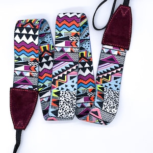 Pode incluir: Uma colorida alça de câmera com um padrão geométrico em tons de roxo, laranja, azul e preto. A alça tem extremidades de camurça bordô e conectores de nylon preto. Projetada para ser usada ao redor do pescoço.