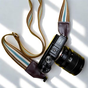 Puede incluir: Una cámara Fujifilm negra con un objetivo de 18-55mm, que incluye una correa con rayas verticales marrones, blancas y azul claro con extremos de cuero marrón oscuro. El cuerpo de la cámara muestra el logo Fujifilm y varios diales.