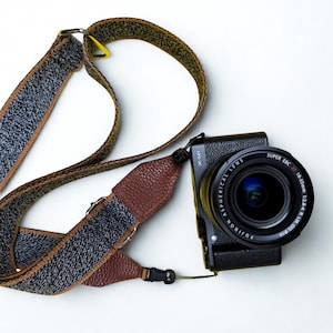 Może przedstawiać: Czarny aparat Fujifilm X-M1 z obiektywem Fujinon Super EBC XF 18-55mm 1:2.8-4 R LM OIS. Aparat jest połączony z długim, regulowanym paskiem o teksturowanym szaro-czarnym wzorze z brązowymi skórzanymi akcentami.