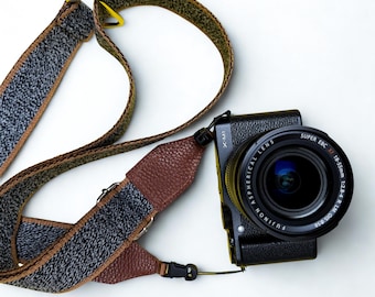 May include: A black digital camera with a Fujinon Super EBC XF 18-55mm lens attached, featuring a textured brown leather and grey speckled fabric camera strap with metal hardware and black plastic buckles, positioned against a plain white background.