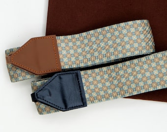May include: Two woven camera straps featuring a repeating geometric pattern in shades of light blue, tan, and cream. Each strap is finished with a pointed end tab, one in smooth brown faux leather with orange stitching and one in black faux leather.