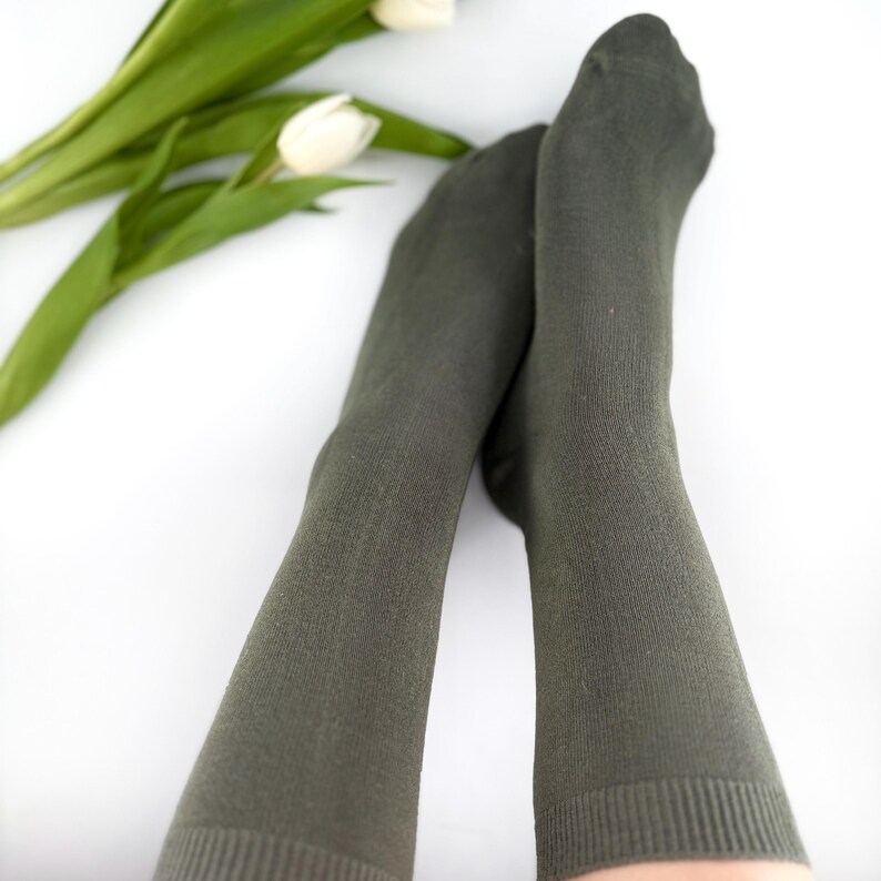 May include: A pair of olive green socks, with a ribbed cuff, are crossed over each other. White tulips with green stems and leaves are in the background. The socks are made of a soft material.