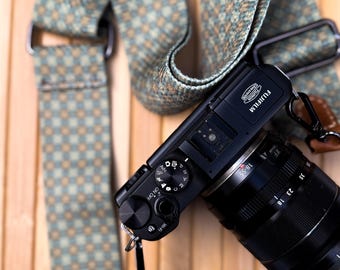 May include: A black Fujifilm camera with an 18-55mm lens attached, featuring a patterned camera strap in shades of teal, tan, and brown. The strap is made of a woven fabric and includes metal hardware for attachment to the camera body.