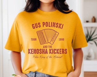 Puede incluir: Una camiseta amarilla con un diseño gráfico rojo que presenta un acordeón y el texto "Gus Polinski and the Kenosha Kickers Polka King of the Midwest!" con los años "1990" y "Tour" a cada lado del acordeón.