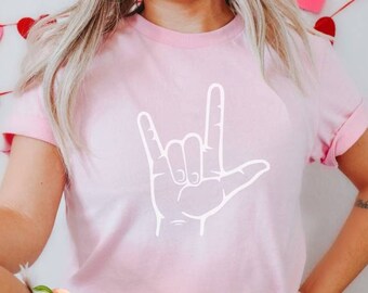 May include: A light pink t-shirt with a white hand gesture graphic, spelling "I Love You" in American Sign Language. The shirt has rolled-up sleeves. Decorative hearts hang in the background. The shirt is a Valentine's Day or romantic theme.