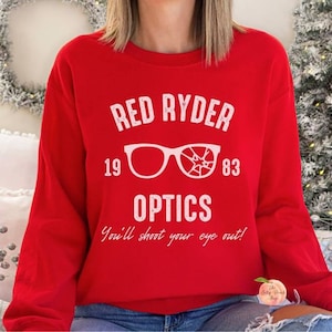May include: A red sweatshirt featuring the text "RED RYDER OPTICS" in white, with the years "19" and "83". The design includes eyeglasses, one intact and one broken. The phrase "You'll shoot your eye out!" is printed below the graphic.