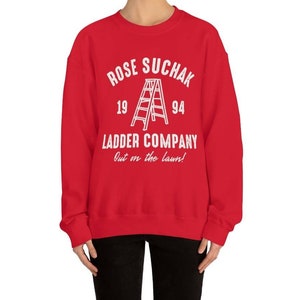 K&ouml;nnte beinhalten: Roter Pullover mit wei&szlig;em Text, der "Rose Suchak Ladder Company Out on the lawn!" lautet, mit einer Leitergrafik und den Zahlen 19 und 94.