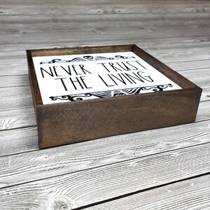 May include: A wooden sign with a white background and black lettering that reads "Never Trust The Living". The sign is framed in a dark brown wood frame.