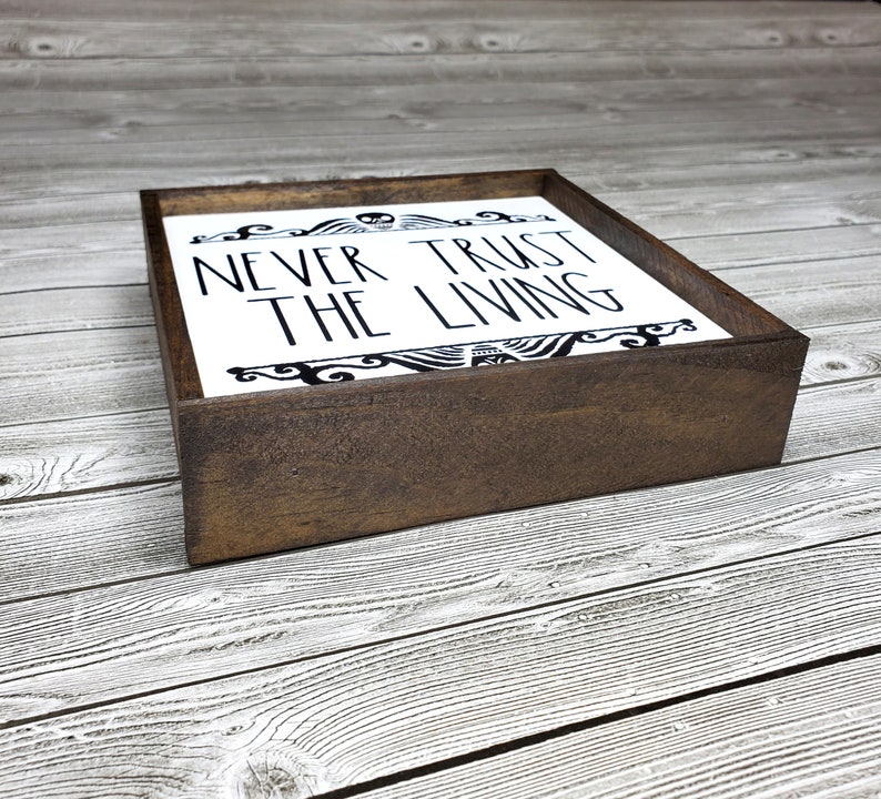 May include: A wooden sign with a white background and black lettering that reads "Never Trust The Living". The sign is framed in a dark brown wood frame.