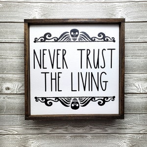 May include: A wooden framed sign with a white background and black text that reads "Never Trust The Living". The sign has a decorative border with a skull and wings at the top and bottom.