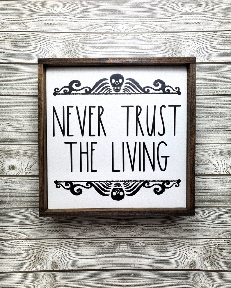 May include: A wooden framed sign with a white background and black text that reads "Never Trust The Living". The sign has a decorative border with a skull and wings at the top and bottom.