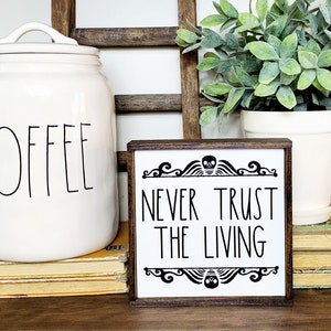 May include: A white wooden sign with black text that reads "Never Trust The Living". The sign has a skull and scroll design at the top and bottom. The sign is sitting on a stack of books.
