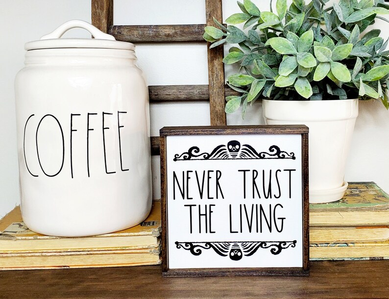 May include: A white wooden sign with black text that reads "Never Trust The Living". The sign has a skull and scroll design at the top and bottom. The sign is sitting on a stack of books.