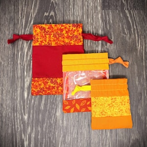 Puede incluir: Tres bolsas con cordón hechas a mano en tonos de rojo, naranja y amarillo. La bolsa más grande es roja con detalles florales naranjas. La bolsa del medio tiene una ventana transparente. La bolsa más pequeña es naranja con detalles florales amarillos.