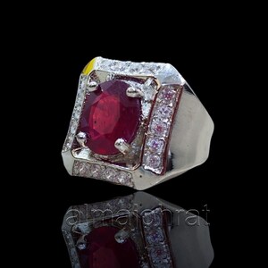May include: A silver ring with a large, oval-cut, deep red gemstone as the centerpiece. The gemstone is surrounded by a square frame of smaller, clear stones. The ring has a wide band and a polished finish, reflecting light.