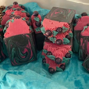 Puede incluir: Una colección de jabones hechos a mano con un diseño de remolino en tonos de negro, rosa y verde. Las barras de jabón están decoradas con pequeñas bayas de color púrpura oscuro y hojas verdes.