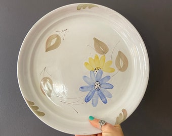 May include: White ceramic plate with a hand-painted floral design. The flowers are blue, yellow, and green. The plate has a raised rim.