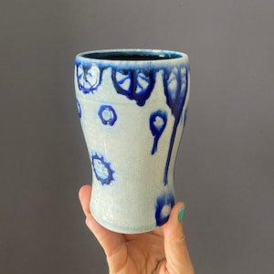 Puede incluir: Una taza de cerámica blanca con un esmalte azul. El esmalte tiene un efecto de goteo y crea un patrón único.