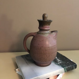 May include: A brown and purple ceramic pitcher with a textured surface and a wooden lid. The pitcher is sitting on top of two books.
