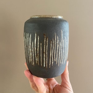 Könnte beinhalten: Eine dunkelgraue Keramikvase mit vertikalen weißen Linien. Die Vase hat eine strukturierte Oberfläche und einen hellbraunen Rand. Die zylindrische Form wird von einer Hand vor einem neutralen Hintergrund gehalten. Diese dekorative Vase eignet sich für die Inneneinrichtung.