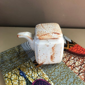 May include: A white ceramic condiment dispenser with a lid and a spout. The dispenser has a rustic, textured finish and is sitting on a colorful fabric.