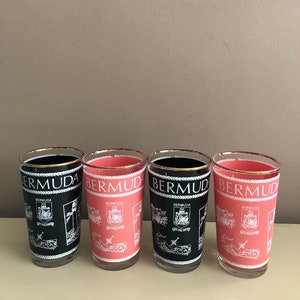 May include: Four glass tumblers with a gold rim and a white design of the Bermuda islands and the Bermuda coat of arms. Two tumblers are black and two are pink.