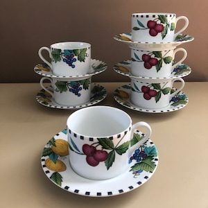 Può includere: Set di sei tazze da tè e piattini in ceramica bianca con un motivo a frutta. Le tazze e i piattini presentano un bordo a scacchi bianco e nero e un design colorato di ciliegie, uva e limoni.