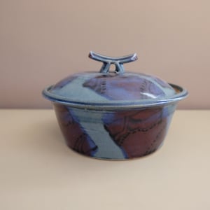 Puede incluir: Una fuente de cerámica hecha a mano con tapa. El plato está pintado con tonos de azul y burdeos. La tapa tiene un pequeño asa. El plato está sobre una superficie blanca.