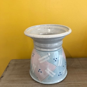 Könnte beinhalten: Eine Keramikvase mit breitem, ausgestelltem Rand und zylindrischem Körper. Die Vase ist mit einem hellblauen, rosa und weißen geometrischen Muster bemalt. Die Vase steht auf einer Holzoberfläche.