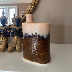 May include: A rectangular ceramic vase with a textured, brown lower half and a light pink upper section. The vase has a narrow neck and a white base. The glaze has a blue and white drip effect where the colors meet.