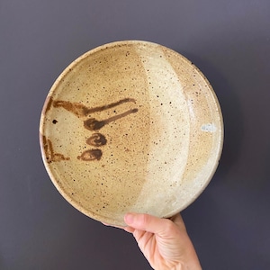 May include: Handmade ceramic plate with a speckled, tan glaze. The plate features a unique, abstract brown design on the left side. The plate is round and has a slightly raised edge. A hand is holding the plate.