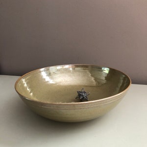 May include: A large, light green ceramic bowl with a slightly iridescent glaze. The bowl has a wide, shallow shape and a slightly rounded rim. A small, silver metal star-shaped object is sitting in the center of the bowl.