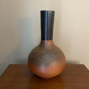 Puede incluir: Un jarrón de cerámica hecho a mano con un cuerpo redondeado y un cuello cilíndrico alto. El jarrón presenta un degradado de colores, pasando de una base marrón rojizo a un cuello marrón oscuro. La superficie tiene una textura moteada.