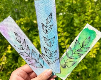 May include: Three watercolor bookmarks with leaf designs. Each bookmark features a different color scheme: purple and pink, blue, and green. The leaf design is a black outline. The bookmarks are held in front of a green, leafy background.