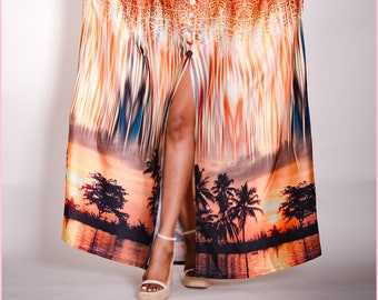 May include: A long, flowing, orange, green, and blue patterned maxi dress with a button-up front and a high slit. The dress has a tropical sunset print with palm trees and a lake.