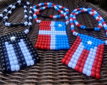 May include: Three beaded necklaces with flag-shaped pendants. The necklaces are made of white and red beads, white and blue beads, and black and white beads. Each pendant features a different flag design with a white star in the center.
